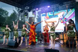 Grupa tancerzy i muzyków w kolorowych strojach podczas występu na scenie.