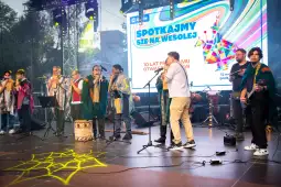 Koncert z zespołem na scenie, kolorowe światła, publiczność w tle.