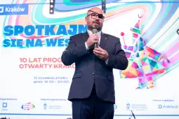 Mężczyzna w okularach przemawia na tle kolorowego plakatu promującego wydarzenie w Krakowie.