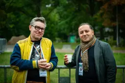 Dwóch mężczyzn w casualowych ubraniach trzymających kubki, stojących na tle zieleni.