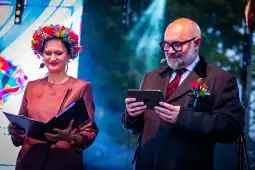 Kobieta w kwiecistej koronie i mężczyzna z tabletem na scenie podczas wydarzenia.