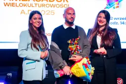 Trzy osoby na scenie, jedna z nagrodą, dwie z bukietami kwiatów.