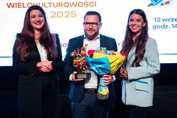 Trzy osoby na scenie z nagrodą i bukietem kwiatów.