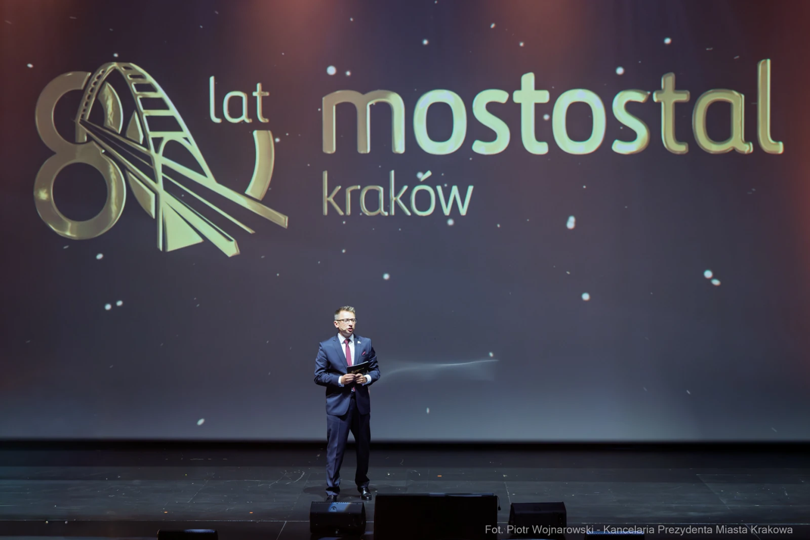 Mężczyzna w garniturze przemawia na scenie z logo Mostostal Kraków z okazji 80-lecia firmy.  Autor: Piotr Wojnarowski