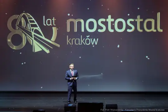Mężczyzna w garniturze przemawia na scenie z logo Mostostal Kraków z okazji 80-lecia firmy.