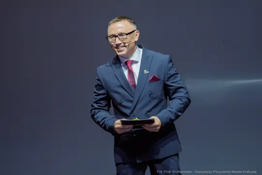Mężczyzna w garniturze z uśmiechem, trzymający mikrofon na tle szarej ściany.