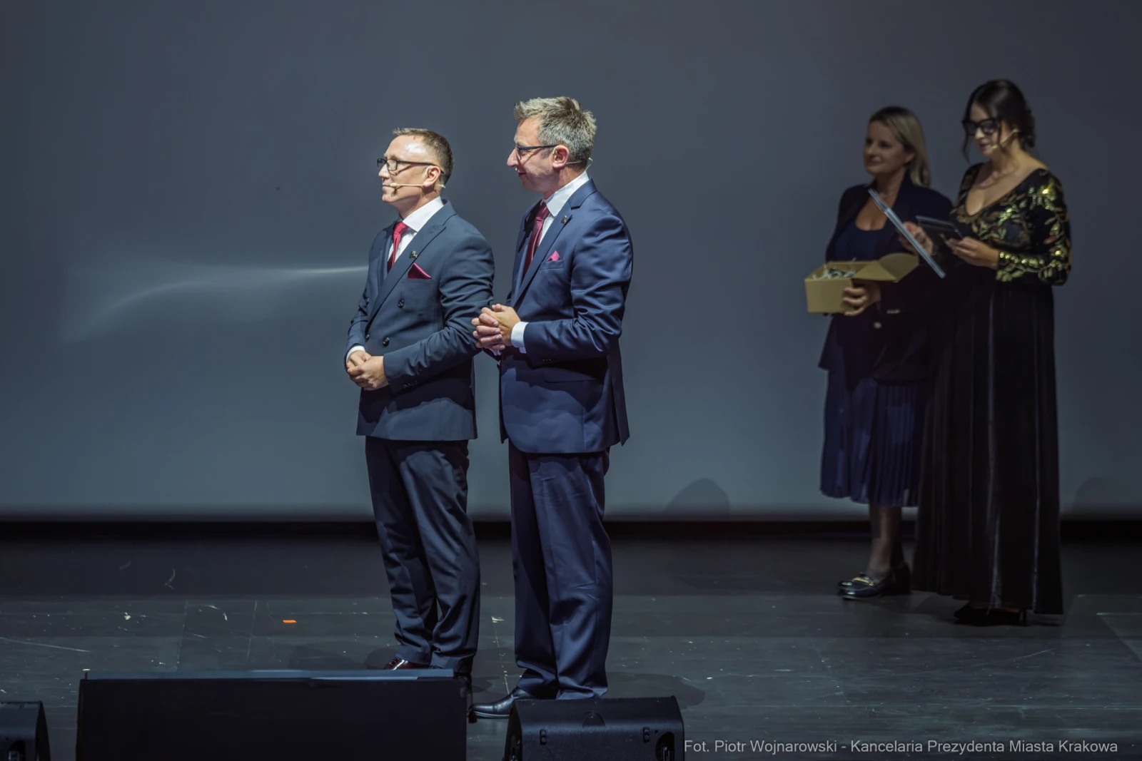 Dwaj mężczyźni w garniturach na scenie, w tle kobiety z nagrodami.  Autor: Piotr Wojnarowski