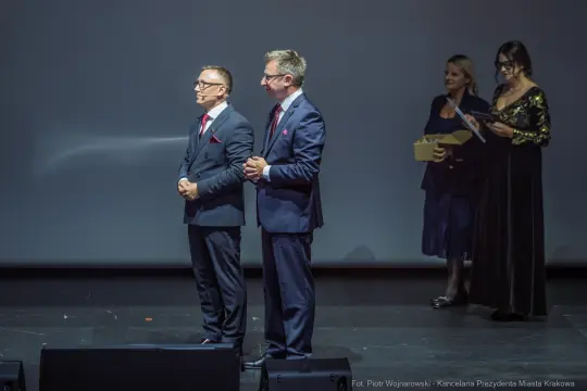 Dwaj mężczyźni w garniturach na scenie, w tle kobiety z nagrodami.