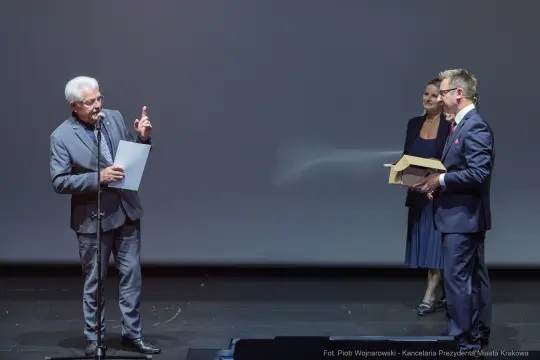 Mężczyzna wręcza nagrodę innemu mężczyźnie na scenie, w tle stoi kobieta.