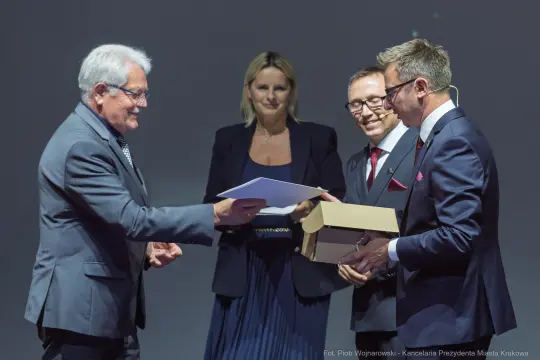 Wręczenie nagrody przez mężczyznę w garniturze, obok dwie osoby w formalnych strojach.