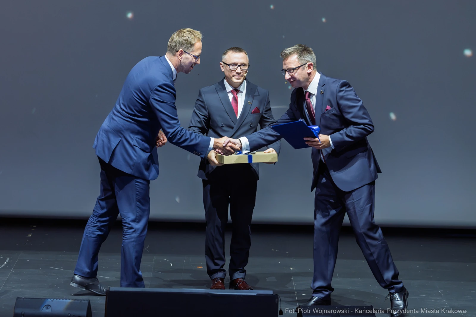 Trzech mężczyzn w garniturach wymienia uściski dłoni i dokumenty na scenie.  Autor: Piotr Wojnarowski