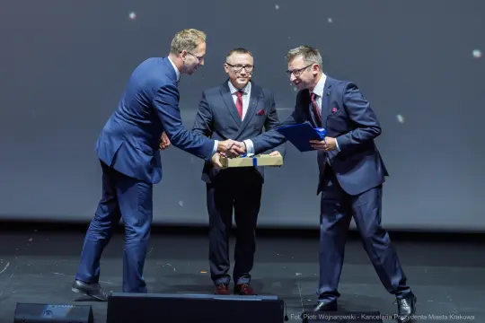 Trzech mężczyzn w garniturach wymienia uściski dłoni i dokumenty na scenie.