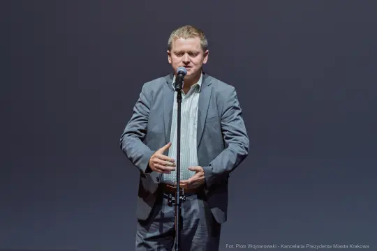 Mężczyzna w garniturze przemawia do mikrofonu na tle ciemnej ściany.