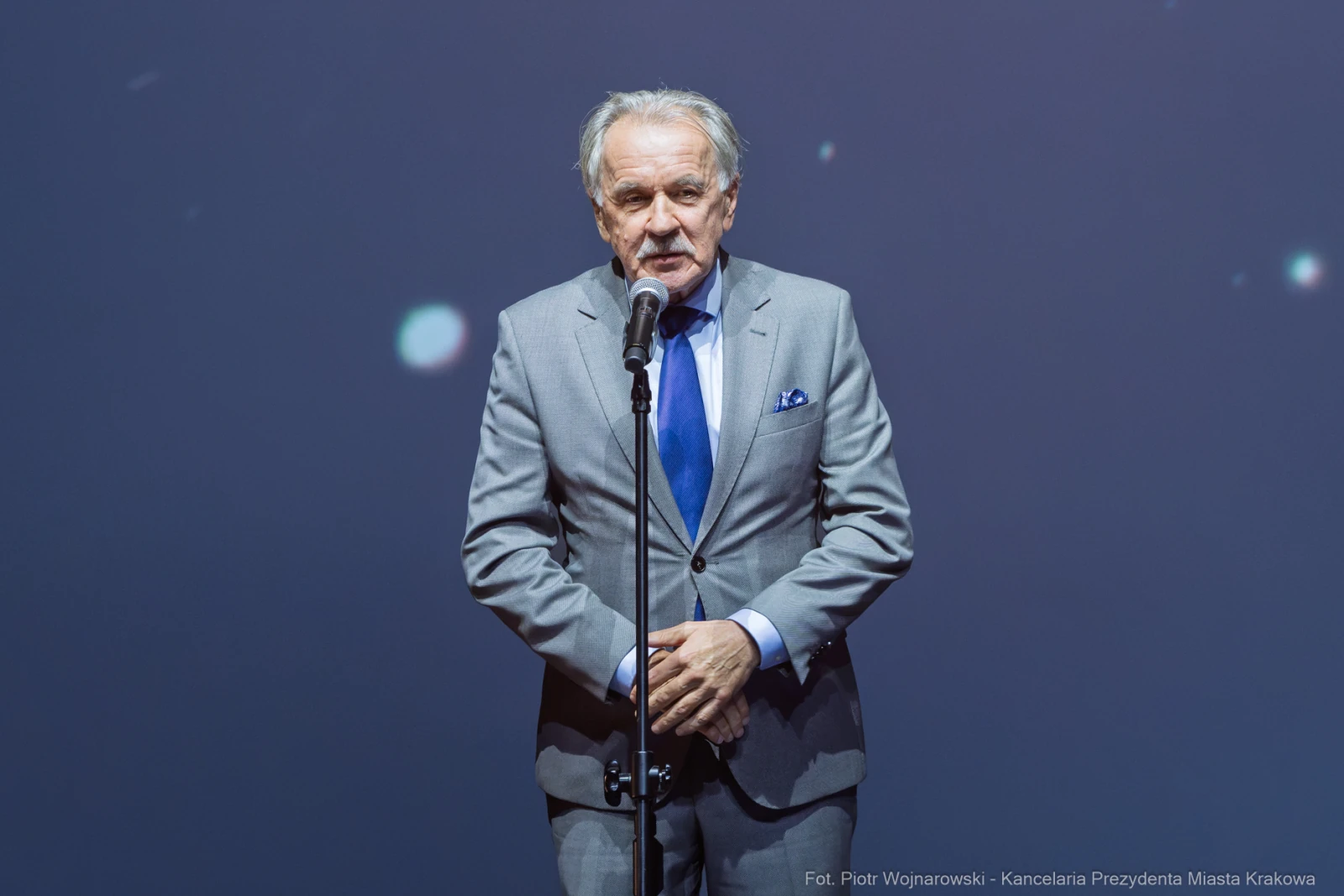 Mężczyzna w szarym garniturze przemawia do mikrofonu na tle ciemnej sceny.  Autor: Piotr Wojnarowski