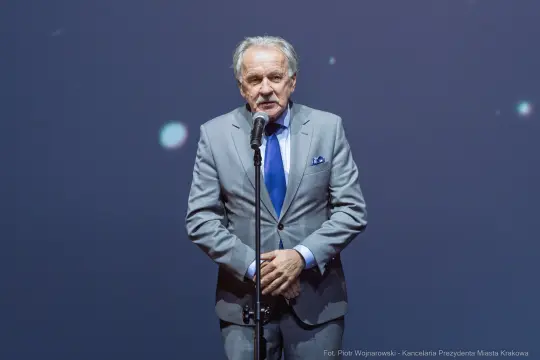 Mężczyzna w szarym garniturze przemawia do mikrofonu na tle ciemnej sceny.