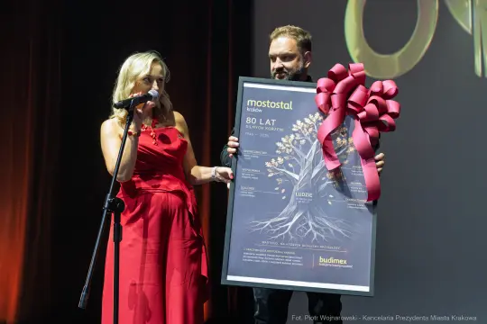 Kobieta w czerwonej sukience wręcza mężczyźnie dyplom z okazji 80-lecia firmy Mostostal.