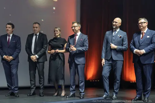 Grupa elegancko ubranych mężczyzn i kobieta na scenie podczas wydarzenia.