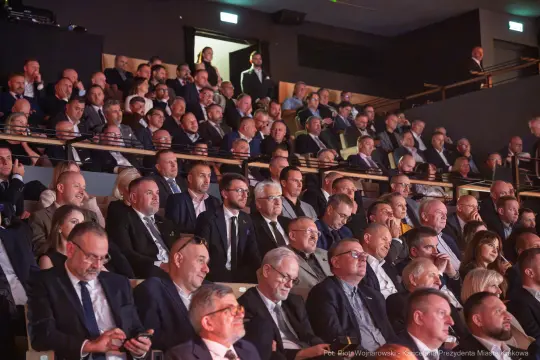 Tłum ludzi w eleganckich strojach na wydarzeniu w sali konferencyjnej.