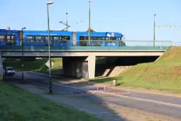 Niebieski tramwaj przejeżdża pod mostem, obok znajduje się droga i zielona trawa.