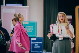 Dwie kobiety w kolorowych ubraniach rozmawiają w tle stoiska wystawowego.