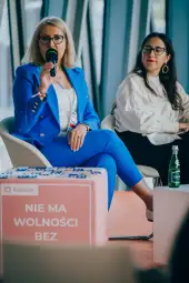 Kobieta w niebieskim garniturze przemawia do mikrofonu, obok siedzi druga kobieta. Na stole napis "NIE MA WOLNOŚCI BEZ".