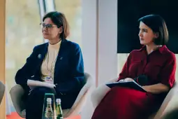 Dwie kobiety siedzące na panelu dyskusyjnym, jedna w niebieskim garniturze, druga w czerwonej sukience.