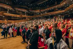 Tłum ludzi na konferencji w nowoczesnej sali, z widocznymi czerwonymi krzesłami.