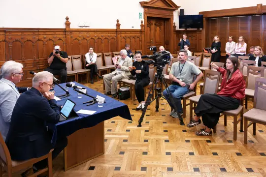 Konferencja prasowa z dziennikarzami i kamerzystami w sali z drewnianymi panelami.
