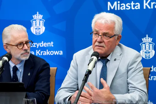 Dwóch mężczyzn podczas konferencji prasowej, jeden mówi do mikrofonu. Tło z logo miasta Krakowa.