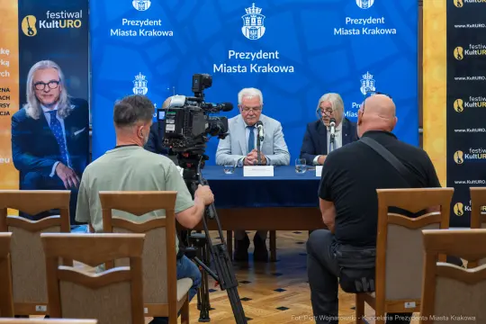 Konferencja prasowa z udziałem dwóch mężczyzn przy stole, kamera i dziennikarze.
