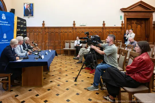 Konferencja prasowa z udziałem kilku mówców i dziennikarzy w sali z drewnianymi detalami.