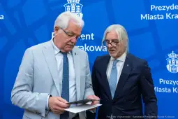 Dwaj mężczyźni w garniturach przeglądają dokumenty na tle niebieskiego tła z logo miasta Krakowa.