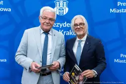 Dwóch mężczyzn w garniturach, trzymających dokumenty, na tle niebieskiego tła z herbem Krakowa.