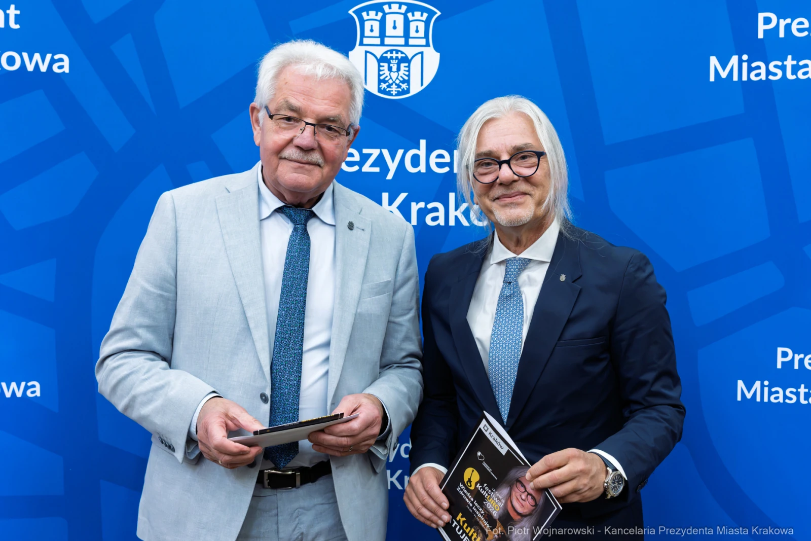 Dwóch mężczyzn w garniturach, trzymających dokumenty, na tle niebieskiego tła z herbem Krakowa.  Autor: Piotr Wojnarowski