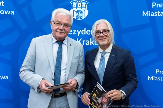Dwóch mężczyzn w garniturach, trzymających dokumenty, na tle niebieskiego tła z herbem Krakowa.