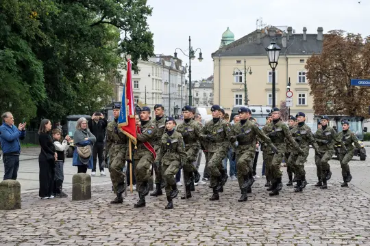 Żołnierze w mundurach maszerują z flagą, obok obserwatorzy na ulicy.