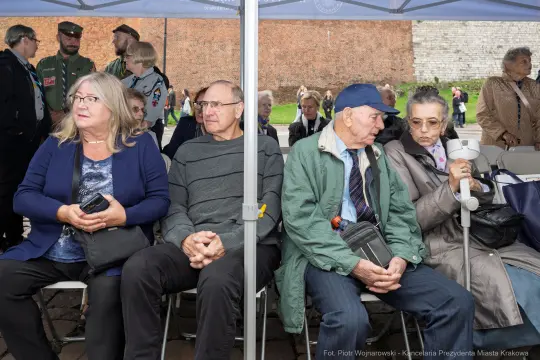 Grupa osób siedzących pod namiotem, w tle widoczni inni uczestnicy wydarzenia.
