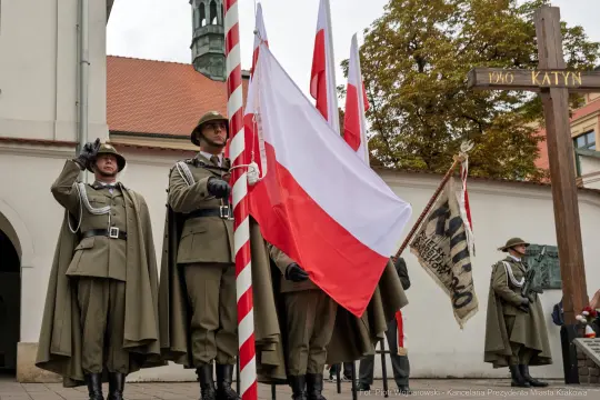 Żołnierze w mundurach trzymają flagę Polski i sztandar, obok krzyż upamiętniający Katyń.