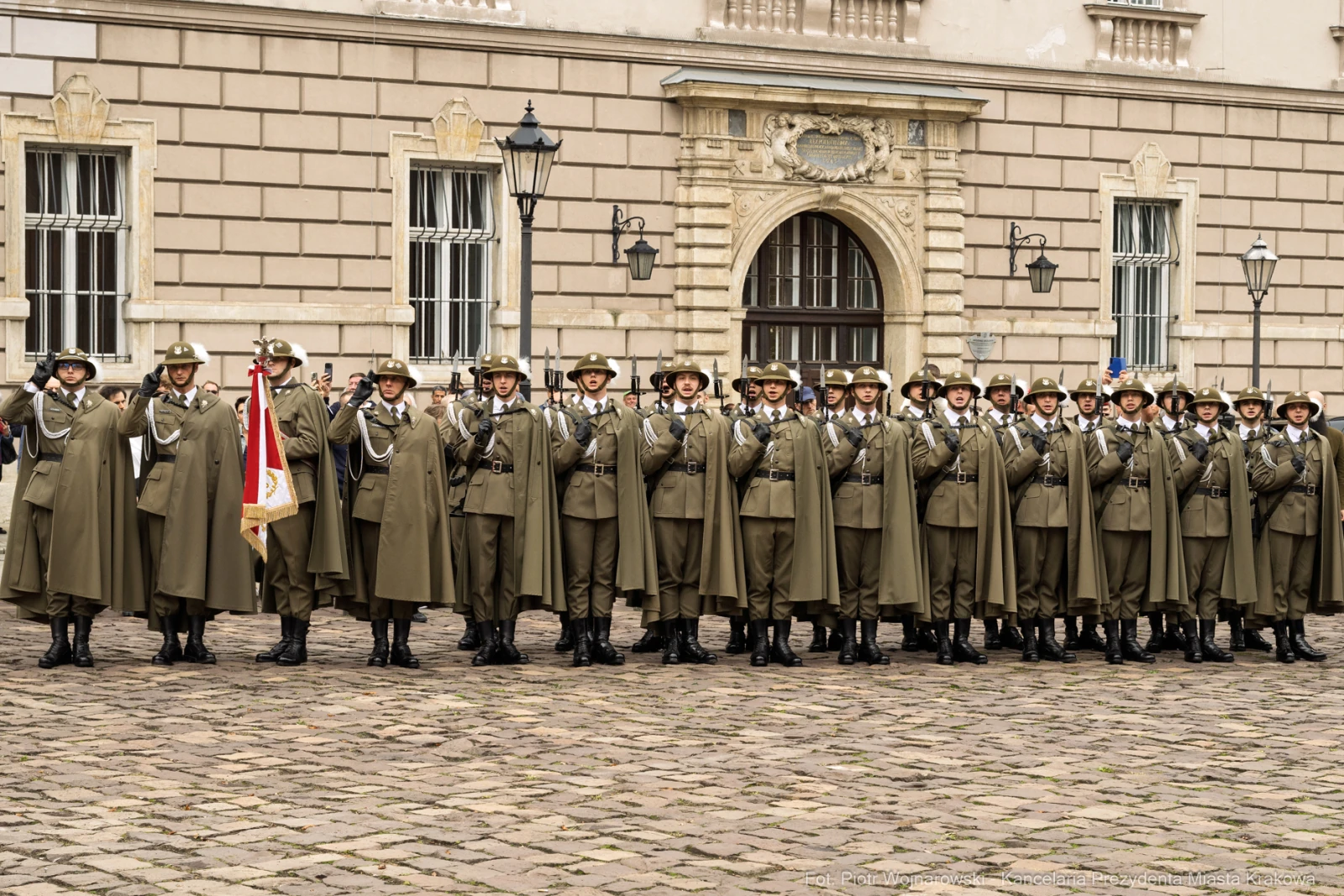 Żołnierze w mundurach stoją w szeregu przed budynkiem, trzymając broń i flagi.  Autor: Piotr Wojnarowski