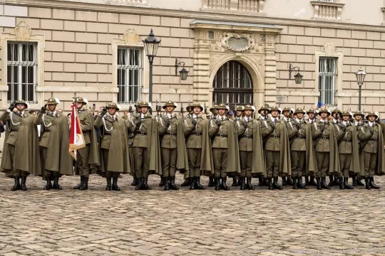 Żołnierze w mundurach stoją w szeregu przed budynkiem, trzymając broń i flagi.