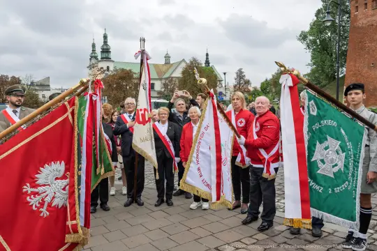 Uczestnicy ceremonii z flagami i sztandarami w historycznym otoczeniu.