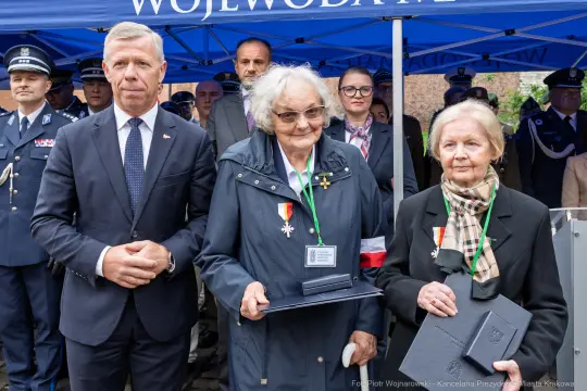 Uroczystość wręczenia odznaczeń dwóm kobietom w obecności mężczyzny w garniturze.