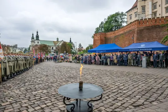 Uroczystość z udziałem wojska, flagi, tłum ludzi i znicz na tle historycznych budowli.