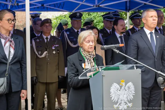 Kobieta przemawia przy mównicy, w tle zgromadzenie osób w mundurach i garniturach.