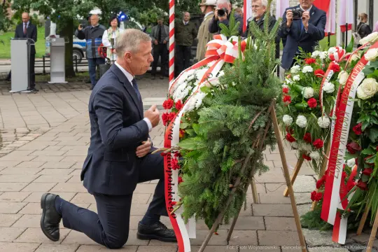 Mężczyzna klęczy przy wieńcu z kwiatów, składając hołd.