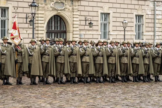 Żołnierze w mundurach stoją w szeregu, trzymając flagę.
