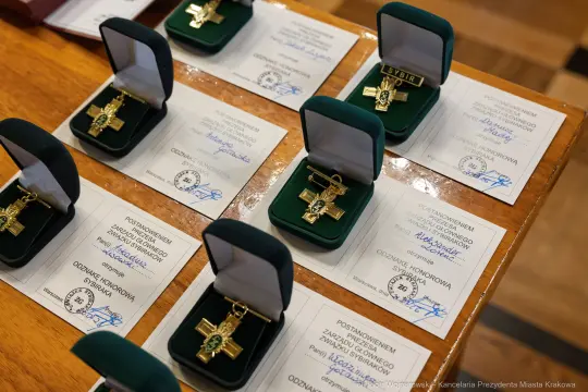 Złote krzyże honorowe w otwartych pudełkach na tle dyplomów.