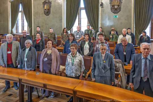 Grupa ludzi stojąca w sali obrad, w tle okna i drewniane stoły.