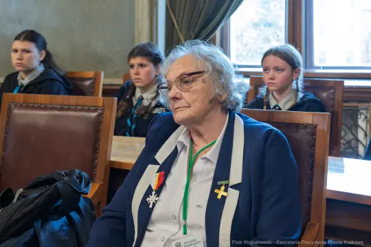 Starsza kobieta w garniturze z odznaczeniem, siedzi w sali obrad, w tle młodzież.