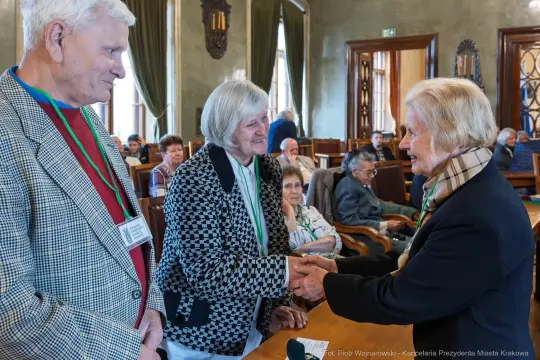 Trzy osoby w rozmowie, jedna z nich podaje rękę drugiej, w tle inni uczestnicy spotkania.